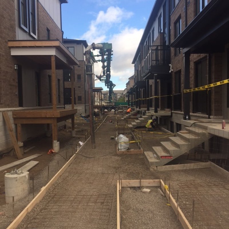 Concrete pump truck operating between residential townhouses with construction workers preparing foundations and site forms under clear sky in Ontario
