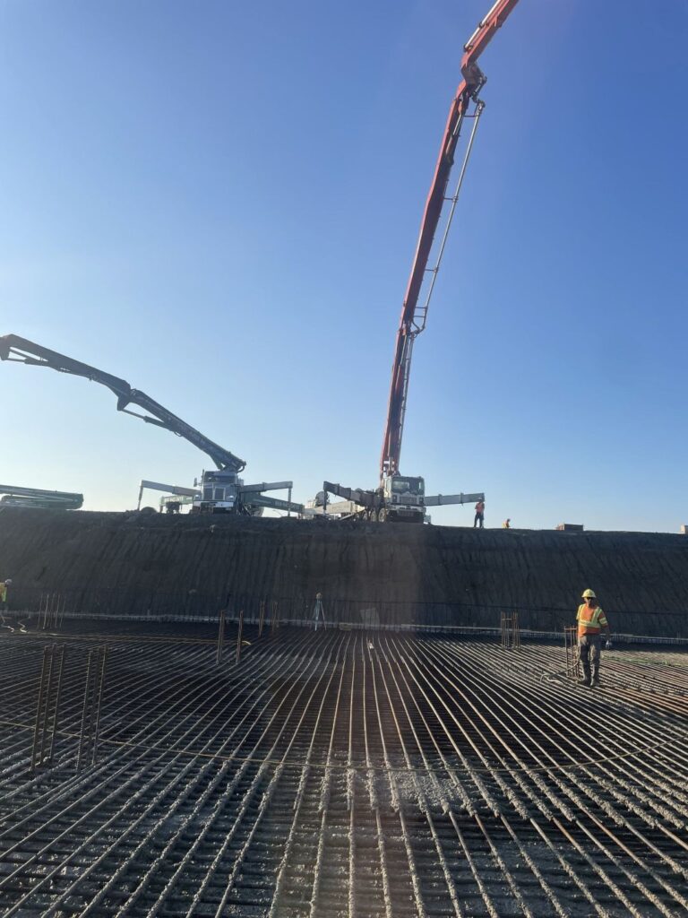 Construction workers pouring concrete over a large rebar grid with a 61m and 50m boom pump on a commercial jobsite, showcasing proper steel reinforcement placement before slab pouring.