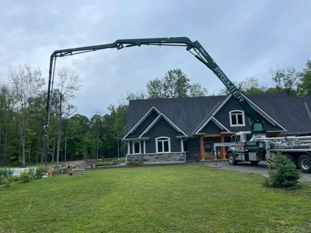 A large 38-meter concrete boom pump is positioned on a residential property, pouring concrete into insulated concrete form (ICF) walls of a newly constructed home surrounded by trees and lawn