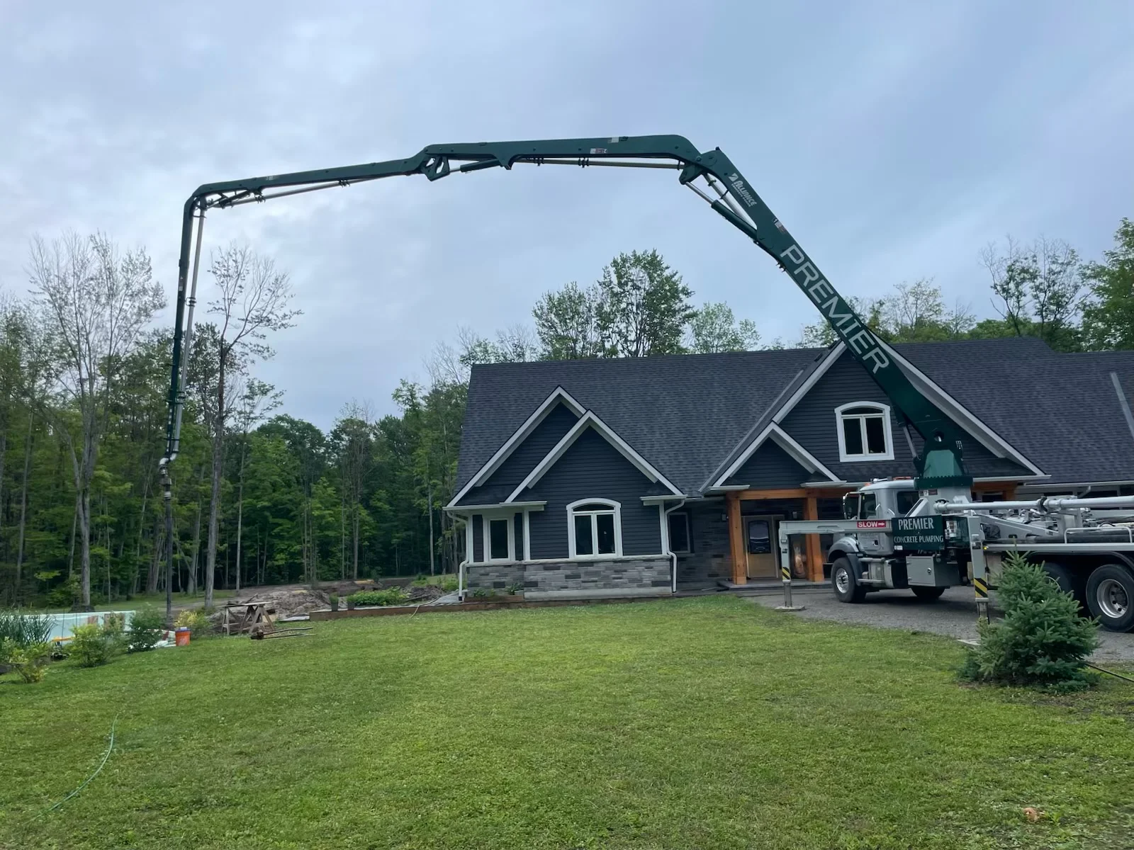 A large 38-meter concrete boom pump is positioned on a residential property, pouring concrete into insulated concrete form (ICF) walls of a newly constructed home surrounded by trees and lawn