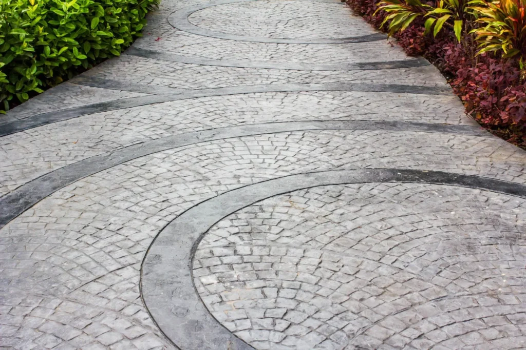 Stamped decorative concrete walkway with intricate curved brick patterns surrounded by landscaped plants.